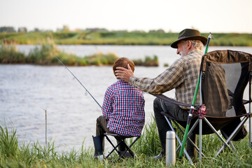 Back view portrait of loving grandfather and boy fishing by lake together during camping trip in...