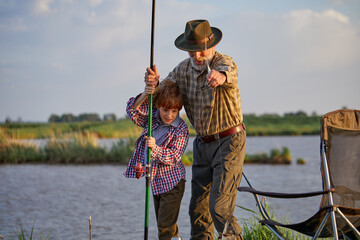 Friendly nice grandfather and grandson pulling rod while fishing on weekend, on like in field, caucasian bearded grandfather help support grandson, wearing casual checkered shirt. side view