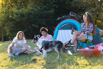 Tres personas de acampada, camping con tienda de campaña. Disfrutando, sonriendo, tocando la guitarra y hablando por teléfono - obrazy, fototapety, plakaty