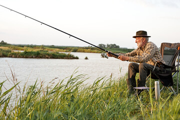 Obraz na płótnie Canvas Side view on elderly gray-haired man fishing on lake in nature, patience and recreation concept.Portrait of senior caucasian fisherman with rod sitting on chair in nature, countryside