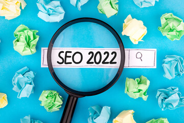 Concept of searching. Text SEO 2022 with magnifier and pieces of paper on blue background.