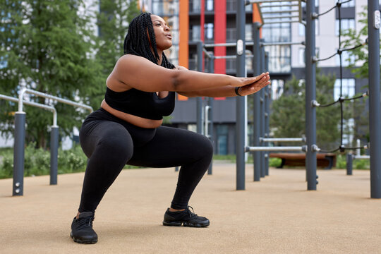 Plus size fat afro american woman in black sportive wear squats in sports ground. Weight losing, obesity, sports, healthcare concept. Side view portrait. Young Woman is concentrated on exercises