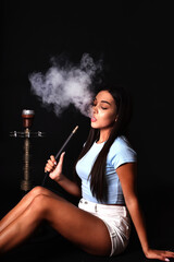 The girl smokes a hookah on a black background. Smoke comes out of the mouth. The pleasure of smoking - obrazy, fototapety, plakaty