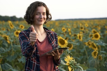 Young joyful woman farmer holding tablet in sunflower field. The use of modern technologies for production of agricultural products. Smiling happy big harvest woman showing positive thumbs up gesture

