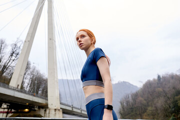 View from down on confident fit redhead female in blue sportswear having rest, taking break after active workout outdoors at the day time, posing, looking forward, ready to run anew. copy space