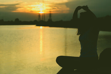 A young girl meditates with her hands raised above her at the lake shore, opposite a beautiful sunset
