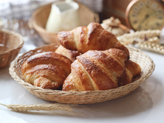 A traditional French croissants in a wooden basket and branch of wheat , with a pot and clock in background . It's a buttery, flaky pastry fold with butter in multiple layers  