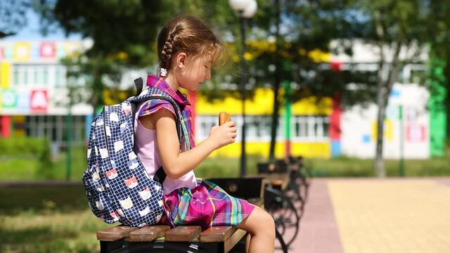 Girl with a backpack sitting on a bench and eating a pie near the school. A quick snack with a bun, unhealthy food, lunch from home. Back to school. Education, primary school classes, September 1