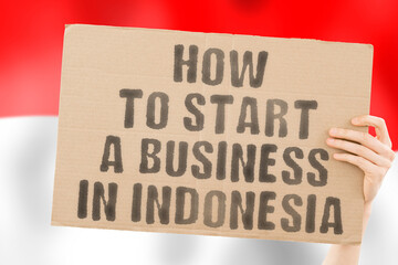 The phrase " How to start a business in Indonesia " on a banner in men's hand with blurred Indonesian flag on the background. Business. Money. Company. Law. Illegal. Lawyer. Earnings. Job