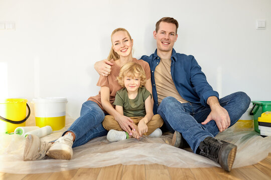 Portrait of happy optimistic family with kid boy sitting on floor with repair tools, at home. Paint buckets next to them. Mother, father and child look at camera, smiling