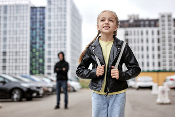 Positive child girl walking alone on street, while terrible maniac looking at her in the background. Focus on kid girl in casual leather jacket enjoy walk, don't afraid of adult men. kidnapping - obrazy, fototapety, plakaty
