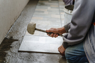 hands of unknown senior man craftsman using hammer to adjust and lay ceramic tiles on the balcony or terrace over adhesive cement in day - construction industry concept copy space