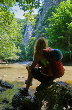 Girl with climbing helmet and a backpack sits on a rock at Varghis gorge, Persani mountains, Romania, near the river and vertical rock walls. Summer adventure.