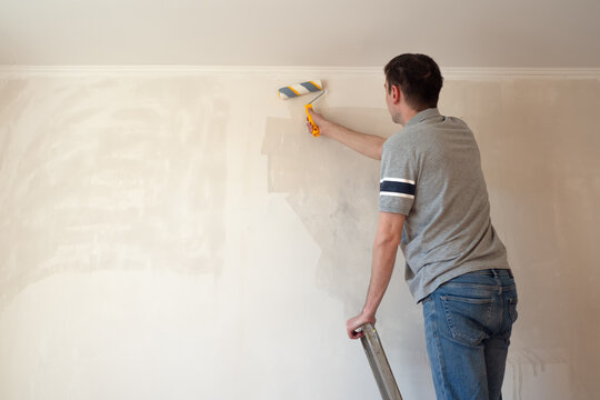 Man priming the wall with a roller. Repair of the interior. Young male decorator painting a wall in the empty room. Concept renovation do it yourself.