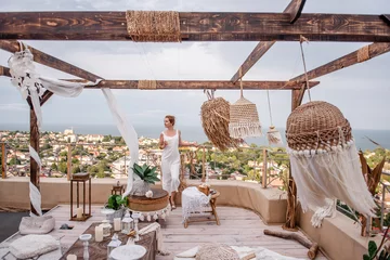 Zelfklevend Fotobehang Young fashion woman in white jumpsuit with styling, holding glass of white wine, stands on the open terrace of the roof of high house overlooking the sea. Stylish girl in Moroccan interior celebrating © farmuty