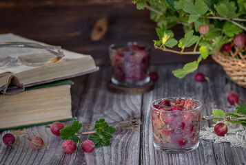 Still life of gooseberry berries on the table with a basket of branches and an open book. Jam in a glass in the foreground.