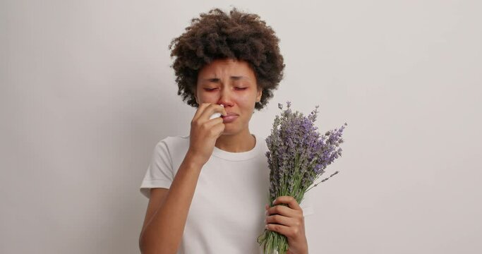 Unhappy curly haired woman suffers from allergy uses nasal spray sneezes all time holds bouquet of lavender has allergic rhinitis red itchy eyes dressed casually isolated over white background