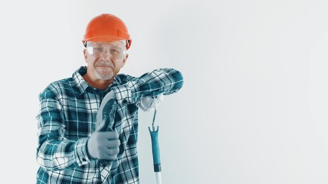 A cheerful gray-haired man in a hard hat and protective glasses holds a paint roller in his hand and smiles happily, having made a good repair in the house. The male artist in the room gives a thumbs