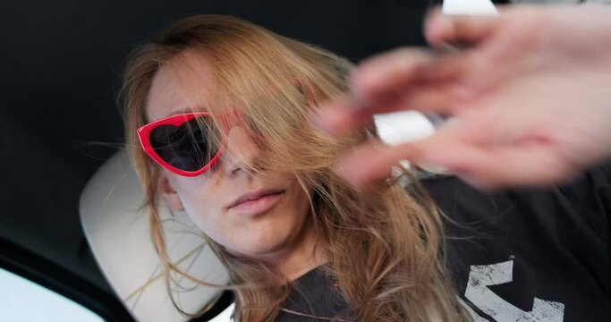 Young happy woman with heart shaped sunglasses making selfie video in car during road trip, showing tongue and goofing around.