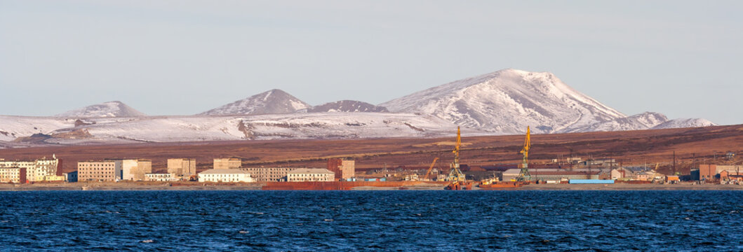 Panorama of the northern port town on the sea coast. In the distance tundra and mountains. Old abandoned buildings. Port cranes of the operating cargo seaport. Beringovsky, Chukotka, Far East Russia.