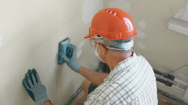 An elderly man master levels the wall with a tool in an orange helmet and protective glasses and a mask, making repairs in a small room.
