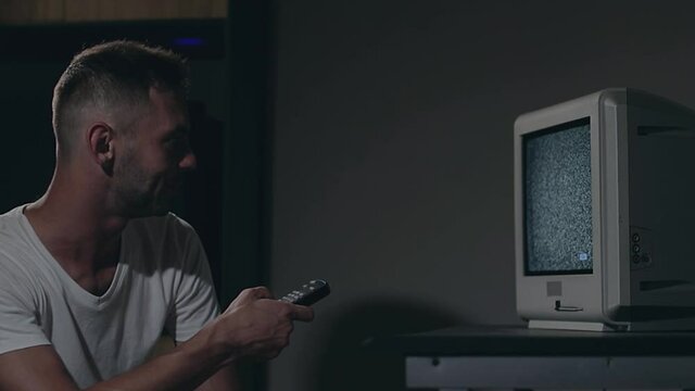 Broken Television or TV Propaganda. Mind Control - Zombified Man in White T-shirt Switches Channels on the Old TV in Dark Room. TV Signal Search. Distortion on the Screen. Medium Close-Up, Film Grain