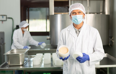 Skilled cheesemaker wearing white robe and protective face mask working in shaping workshop of cheese factory