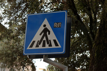 Pedestrian crossing traffic sign, blue background, simple person icon walking on zebra stripes, road safety, urban area, street, transportation, trees in background,  vertical orientation.