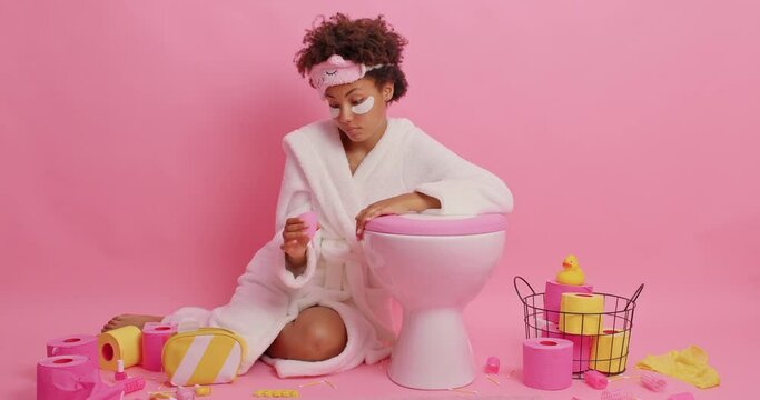Curly haired woman looks attentively on silicone menstruation cup finds better alternative wears sleepmask white bathrobe poses in lavatory near toilet bowl applies patches under eyes. Womans periods