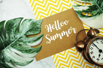 Hello Summer typography text on paper card with alarm clock and Monstera leave