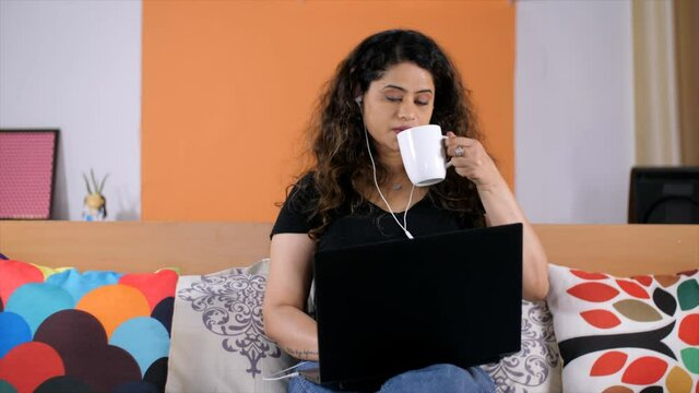 A young freelancer with white earphones working on her laptop - remote job. Pretty Indian female drinking tea / coffee - leisure and relaxation. Work from home  Modern lifestyle