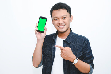 Young Asian man is standing and smiling pointing on green screen on smartphone screen on white background.