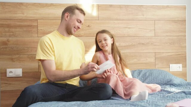Happy daddy reading card given gy little daughter on Father Day, home interior. caucasian family sit on bed having rest, relax at weekends, holidays