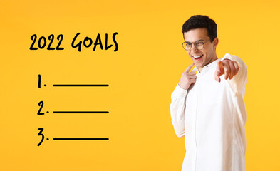 Handsome young man pointing at viewer on yellow background with empty to do list for 2022 year