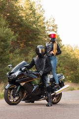 Fototapeta na wymiar Motorcyclist in a leather jacket sits on a motorcycle and a girl stands on a motorcycle in helmets against the background of trees in the forest