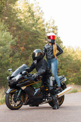 Motorcyclist in a leather jacket sits on a motorcycle and a girl stands on a motorcycle in helmets against the background of trees in the forest