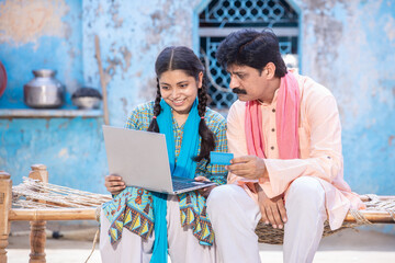Rural Indian father and young daughter using laptop while sitting on traditional wooden bed outside their house, Happy family looking at computer screen. web surfing. Digital india concept.