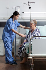Medical nurse helping senior woman patient getting up from bed in hospital room, with iv drip bag...