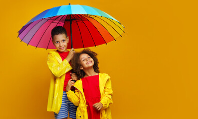 Cheerful child girl and a boy with yellow raincoat under a colored umbrella on colored yellow background.