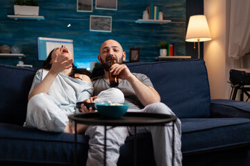 Relaxed couple in pajamas relaxing on sofa eating popcorn watching comedy movie, enjoying free time together. Husband and wife relaxing late at night in living room. Looking at entertainment film