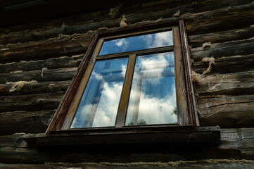 the window of an old rustic log house with a reflection of the blue sky and clouds.