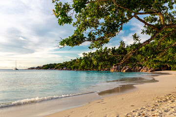 Anse Lazio beach with catamaran anchored in the distance, turquoise water, waves crushing onto sandy beach, tropical vegetation around.
