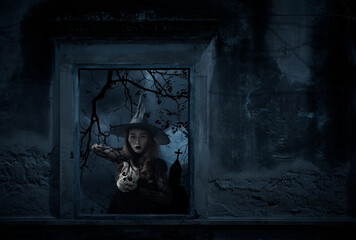 Halloween witch holding a skull standing in old damaged window with wall over cross, church, birds, dead tree and spooky cloudy sky, Halloween mystery concept
