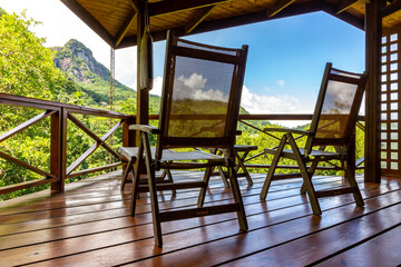 Two wooden chairs on a wooden terrace overlooking Morne Seychelles National Park rainforest with lush tropical vegetation, palm trees and Morne Blanc peak, Mahe Island, Seychelles.