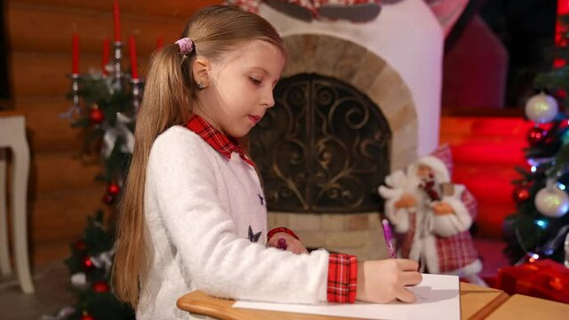 Little girl drawing or writing letter for Santa at Christmas decorated room. Cute child sitting on floor and creating picture on small desks. Christmas mood concept