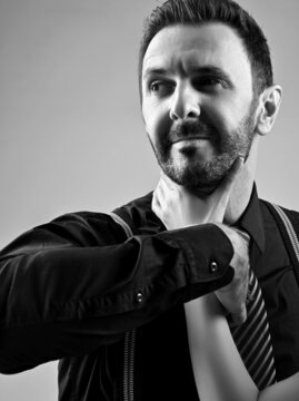 Black and white portrait of brutal bearded man in black shirt with striped tie and suspenders, removing woman hand that was holding him by the throat over light grey background