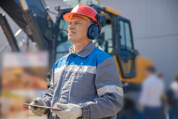 Industrial worker male senior in helmet holds tablet computer on background of production of...