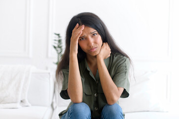 Unhappy young indian woman sitting on sofa, covered forehead with hand and feel frustration, tired...