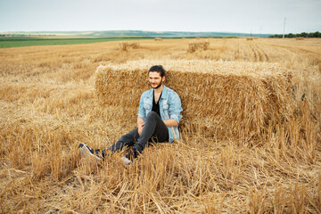 Naklejka na ściany i meble Outdoor photo of young guy in denim jacket, sitting in the wheat field.