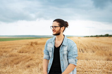Young smiling man in denim jacket standing in farm field near haystacks.